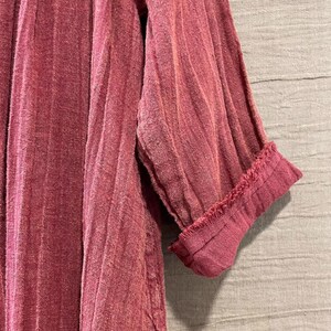 May include: Close-up of a dusty rose-colored garment with vertical ribbed texture. The sleeve is cuffed, revealing a frayed edge. The fabric appears to be lightweight and textured, suggesting a casual style.