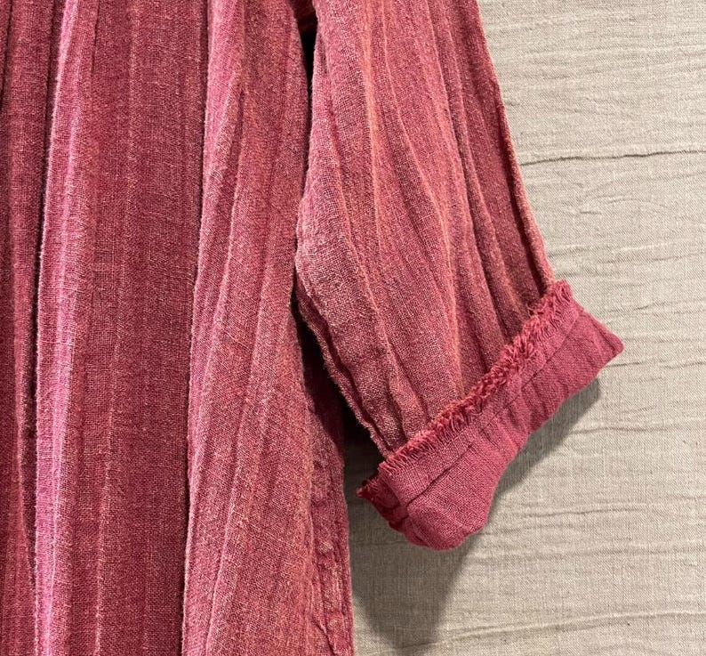 May include: Close-up of a dusty rose-colored garment with vertical ribbed texture. The sleeve is cuffed, revealing a frayed edge. The fabric appears to be lightweight and textured, suggesting a casual style.