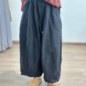 May include: Black linen pants with a wide, flowing silhouette. The pants have a high waist and are paired with tan slip-on shoes. The model is wearing a rust-colored long-sleeve shirt. The pants appear to be made of a lightweight fabric.