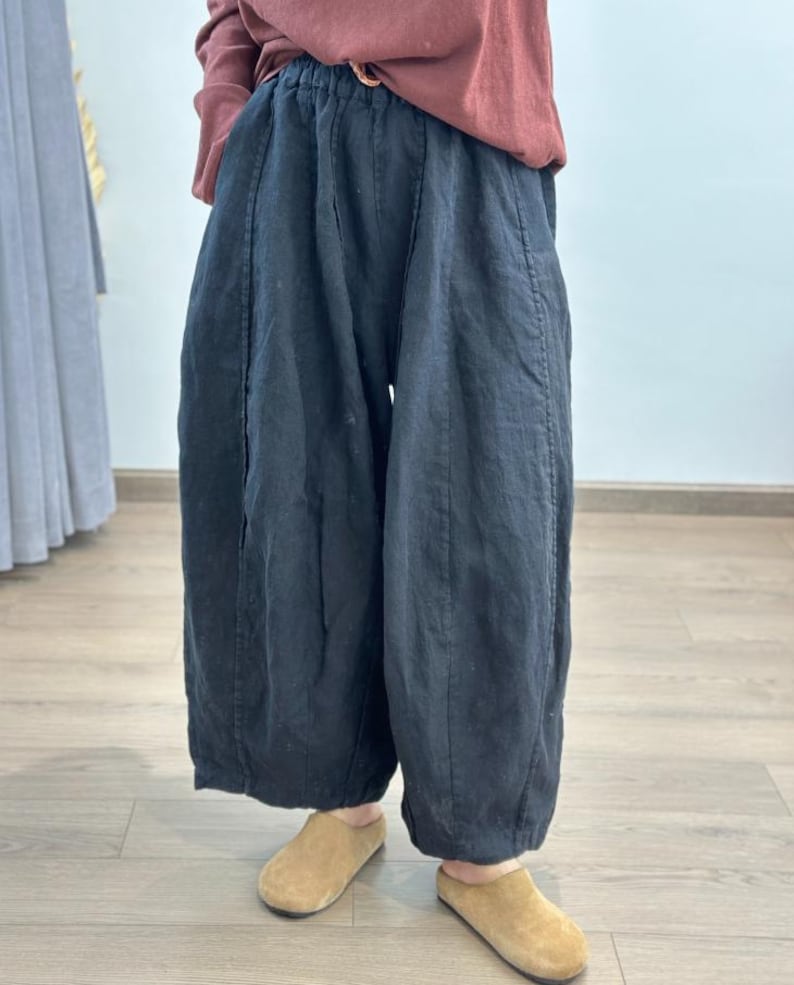 May include: Black linen pants with a wide, flowing silhouette. The pants have a high waist and are paired with tan slip-on shoes. The model is wearing a rust-colored long-sleeve shirt. The pants appear to be made of a lightweight fabric.