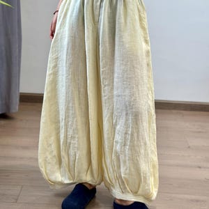 May include: Pale yellow linen pants with a loose, flowing design. The pants have a button closure and pockets. The pants are paired with navy blue slip-on shoes. The pants are a light, neutral color.