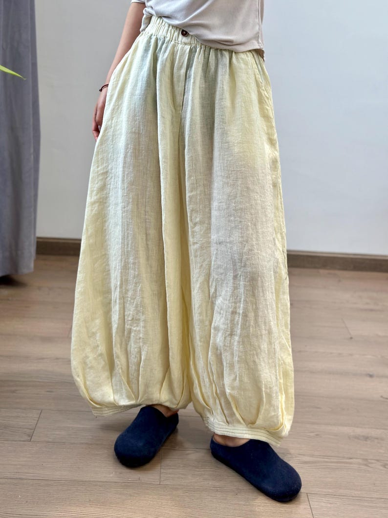May include: Pale yellow linen pants with a loose, flowing design. The pants have a button closure and pockets. The pants are paired with navy blue slip-on shoes. The pants are a light, neutral color.