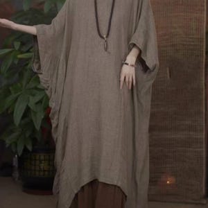 May include: A loose-fitting, taupe-colored tunic dress with wide, batwing-style sleeves and a rounded neckline. The garment features a textured fabric and is layered over dark brown wide-leg trousers. A long beaded necklace with a pendant is worn.