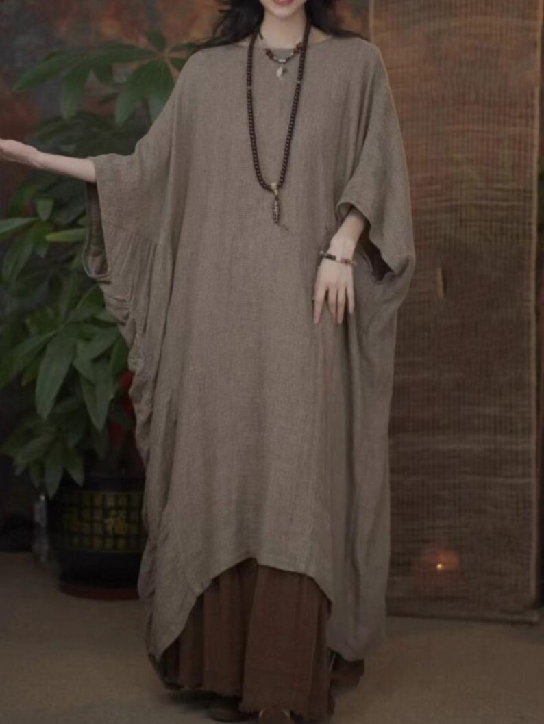 May include: A loose-fitting, taupe-colored tunic dress with wide, batwing-style sleeves and a rounded neckline. The garment features a textured fabric and is layered over dark brown wide-leg trousers. A long beaded necklace with a pendant is worn.