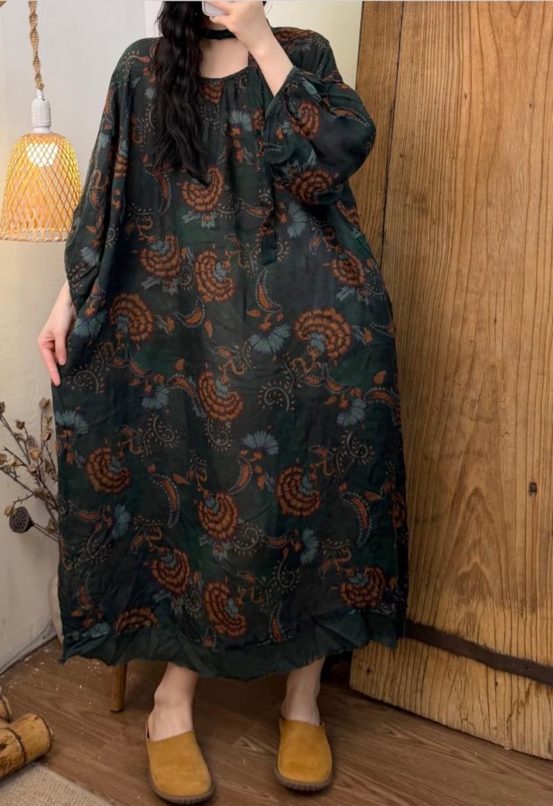 May include: Dark green, long-sleeve dress with a floral paisley pattern in shades of orange and blue. The dress has a loose, flowing silhouette and a round neckline. The dress is paired with brown slip-on shoes.