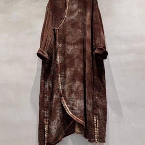 May include: A long, brown, wrap-style coat with a mandarin collar and long sleeves. The coat features a textured fabric with lighter brown accents and a contrasting trim along the edges. The coat has a unique asymmetrical design and is hanging on a wooden hanger.