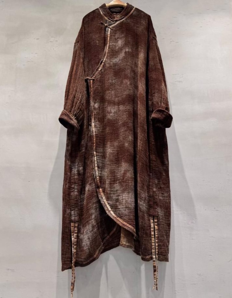 May include: A long, brown, wrap-style coat with a mandarin collar and long sleeves. The coat features a textured fabric with lighter brown accents and a contrasting trim along the edges. The coat has a unique asymmetrical design and is hanging on a wooden hanger.