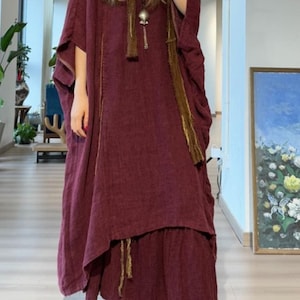 May include: A burgundy-colored, loose-fitting dress with wide sleeves and a layered skirt. The dress features gold-colored trim and a matching scarf with a pendant. The model is wearing beige slip-on shoes. The background includes a painting and indoor plants.