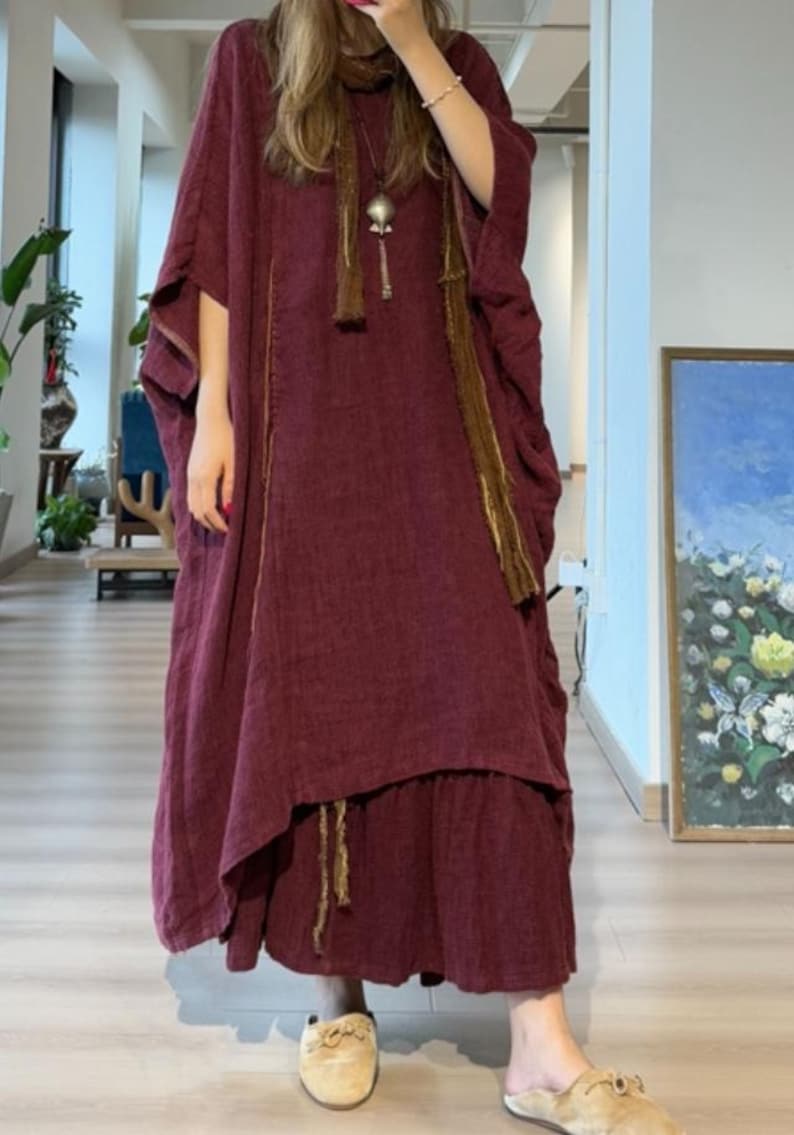 May include: A burgundy-colored, loose-fitting dress with wide sleeves and a layered skirt. The dress features gold-colored trim and a matching scarf with a pendant. The model is wearing beige slip-on shoes. The background includes a painting and indoor plants.