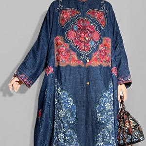 May include: A long, dark blue denim coat with intricate floral embroidery in red, pink, and brown. The coat features a mandarin collar, button closures, and decorative patterns along the hem and sleeves. A matching floral handbag is held in the hand.