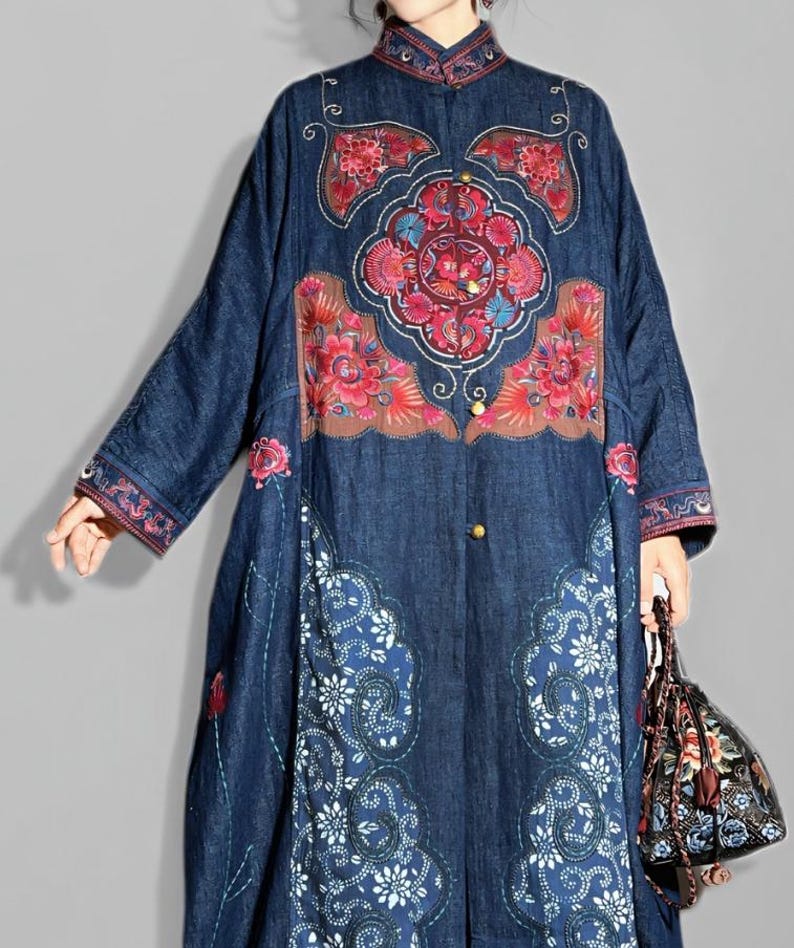 May include: A long, dark blue denim coat with intricate floral embroidery in red, pink, and brown. The coat features a mandarin collar, button closures, and decorative patterns along the hem and sleeves. A matching floral handbag is held in the hand.