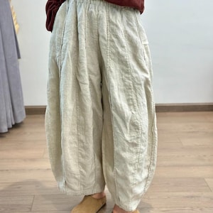 May include: Cream-colored linen pants with a unique, voluminous silhouette. The pants feature vertical stitching details and a gathered ankle. Paired with brown suede slip-on shoes. The pants have a relaxed, comfortable style.