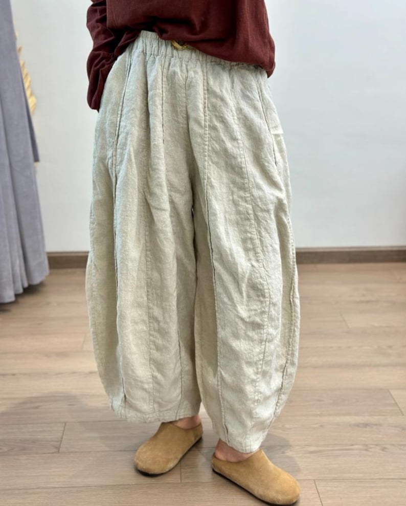 May include: Cream-colored linen pants with a unique, voluminous silhouette. The pants feature vertical stitching details and a gathered ankle. Paired with brown suede slip-on shoes. The pants have a relaxed, comfortable style.