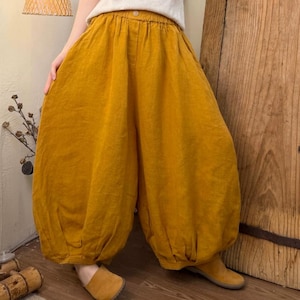 May include: Mustard yellow linen pants with a wide, voluminous silhouette. The pants have a high waist and a button closure. The fabric appears soft and flowing. The pants are paired with a white top and tan shoes.