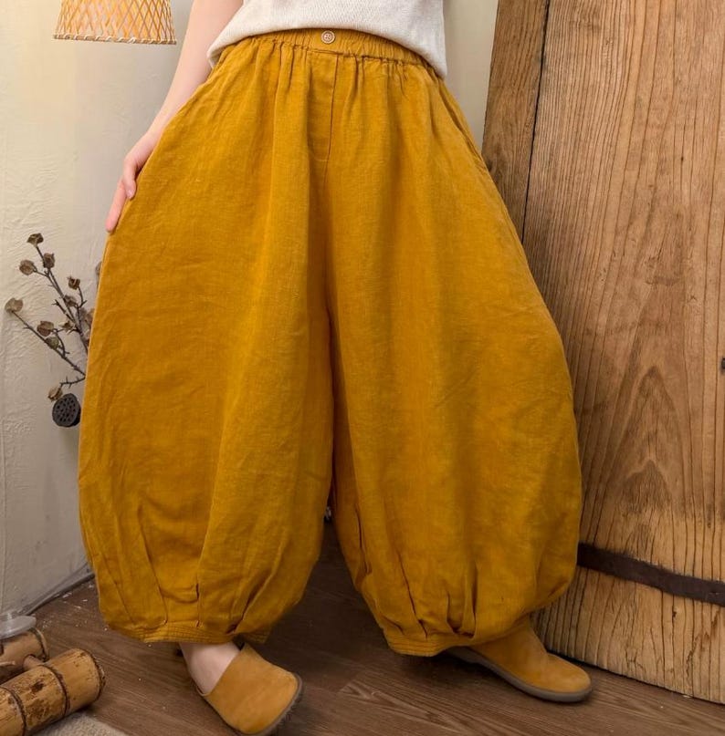 May include: Mustard yellow linen pants with a wide, voluminous silhouette. The pants have a high waist and a button closure. The fabric appears soft and flowing. The pants are paired with a white top and tan shoes.
