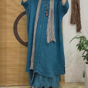 May include: A teal blue linen dress with a long, flowing skirt and a relaxed fit. The dress has a simple design with a round neckline and three-quarter sleeves. The model is wearing a gray scarf and a beaded necklace.