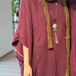 May include: A burgundy-colored, loose-fitting tunic with wide sleeves. A brown scarf is draped around the neck, and a silver pendant necklace hangs down the front. The tunic has a textured appearance and a stitched seam detail.