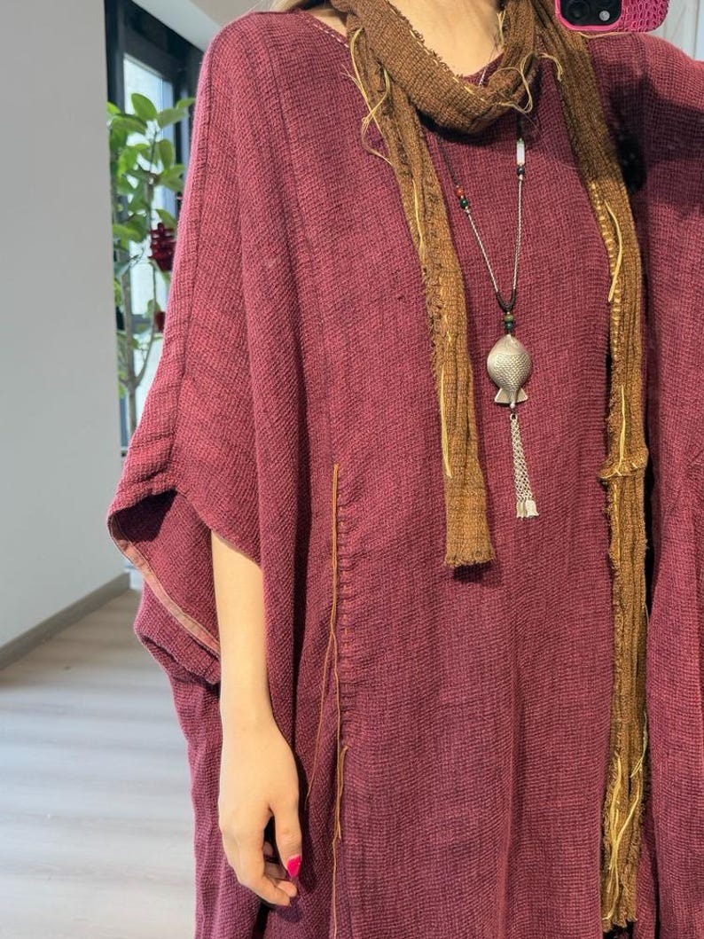 May include: A burgundy-colored, loose-fitting tunic with wide sleeves. A brown scarf is draped around the neck, and a silver pendant necklace hangs down the front. The tunic has a textured appearance and a stitched seam detail.