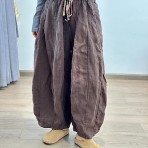 May include: Brown linen pants with a drawstring waist and a loose, flowing design. The pants have a wide leg and are gathered at the ankle. The drawstring is multi-colored. The person is wearing tan colored shoes.