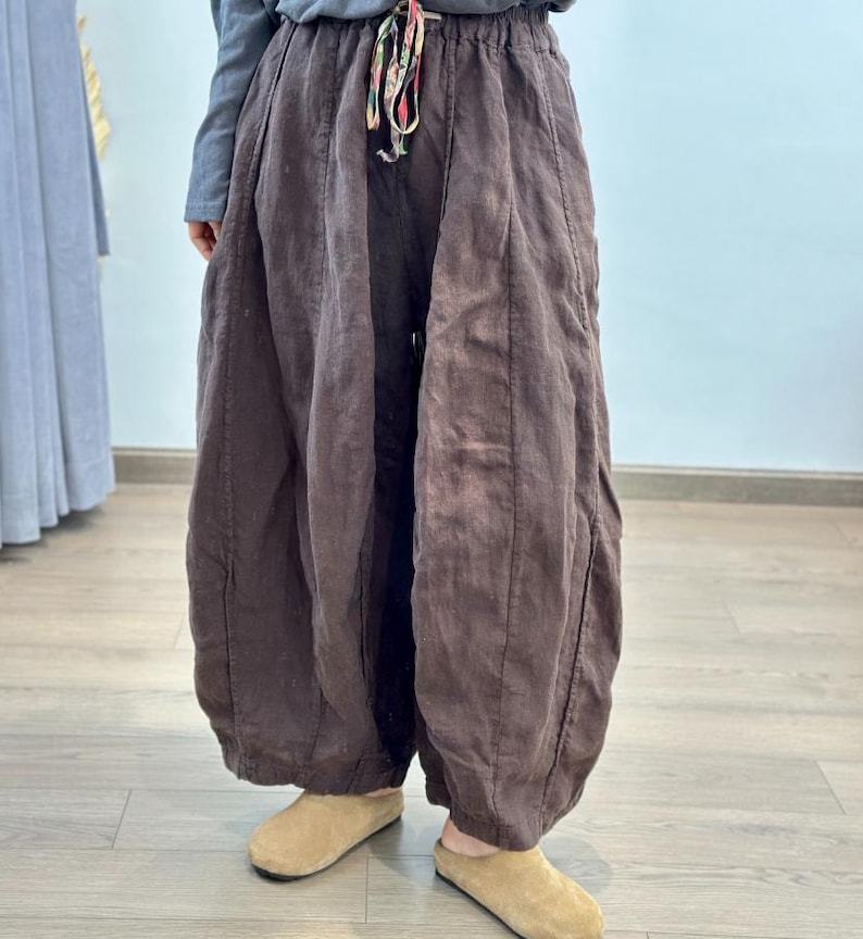 May include: Brown linen pants with a drawstring waist and a loose, flowing design. The pants have a wide leg and are gathered at the ankle. The drawstring is multi-colored. The person is wearing tan colored shoes.