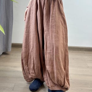 May include: Loose-fitting, brown linen pants with a gathered waist and voluminous, balloon-like legs. The pants are paired with dark blue shoes. The fabric appears lightweight and breathable, suggesting a casual, comfortable style.
