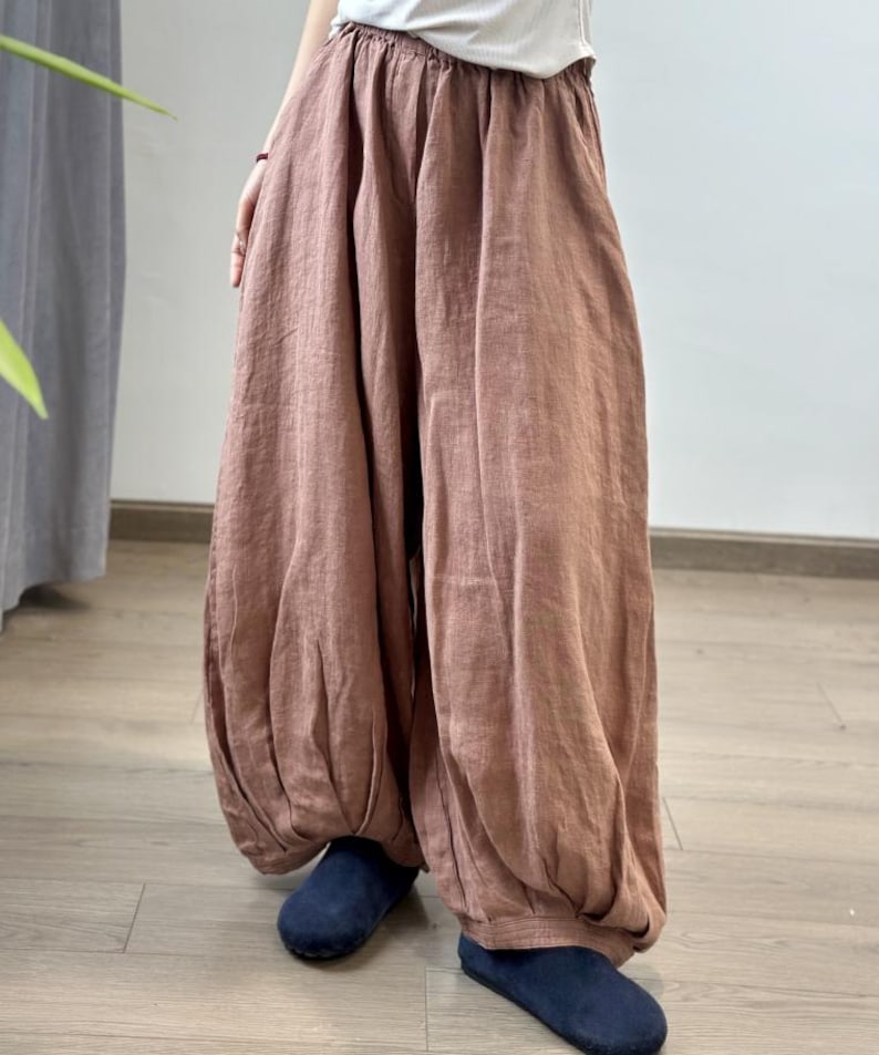 May include: Loose-fitting, brown linen pants with a gathered waist and voluminous, balloon-like legs. The pants are paired with dark blue shoes. The fabric appears lightweight and breathable, suggesting a casual, comfortable style.