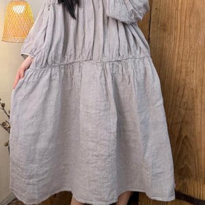 May include: A light gray linen dress with a V-neck and long, puffy sleeves. The dress has a tiered skirt and falls to the mid-calf. The model is wearing yellow-brown slip-on shoes. The background includes a wooden door and a hanging lamp.
