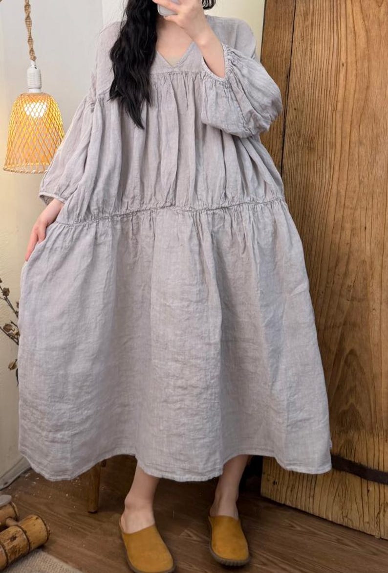 May include: A light gray linen dress with a V-neck and long, puffy sleeves. The dress has a tiered skirt and falls to the mid-calf. The model is wearing yellow-brown slip-on shoes. The background includes a wooden door and a hanging lamp.