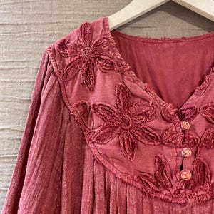 May include: A long-sleeved, dusty rose-colored top with a V-neckline. The top features embroidered floral appliques around the neckline and three small buttons. The fabric has a textured appearance, and the garment hangs on a wooden hanger.
