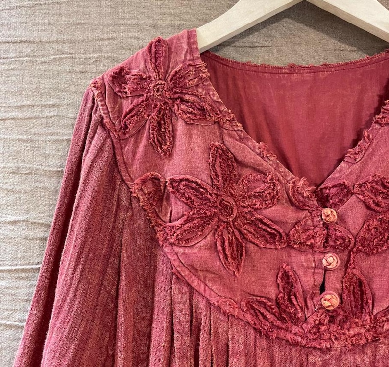 May include: A long-sleeved, dusty rose-colored top with a V-neckline. The top features embroidered floral appliques around the neckline and three small buttons. The fabric has a textured appearance, and the garment hangs on a wooden hanger.