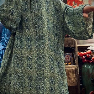 May include: A loose-fitting tunic featuring a repeating intricate damask pattern in shades of sage green, olive, and deep navy blue. The garment has a round neckline and wide, three-quarter length sleeves, providing a relaxed silhouette.