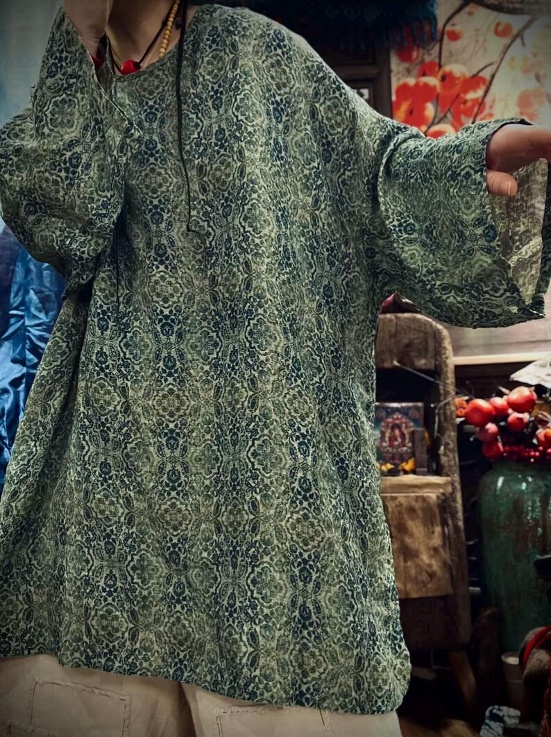 May include: A loose-fitting tunic featuring a repeating intricate damask pattern in shades of sage green, olive, and deep navy blue. The garment has a round neckline and wide, three-quarter length sleeves, providing a relaxed silhouette.