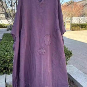 May include: A plum-coloured linen dress with a V-neck and short sleeves. The dress features embroidered floral designs and a loose, flowing silhouette. The dress is hanging on a wooden hanger outdoors, suggesting a casual, comfortable style.
