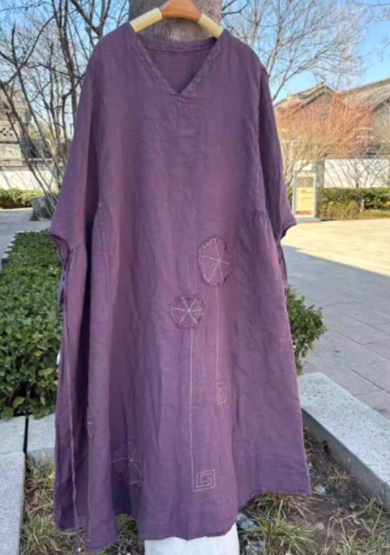 May include: A plum-coloured linen dress with a V-neck and short sleeves. The dress features embroidered floral designs and a loose, flowing silhouette. The dress is hanging on a wooden hanger outdoors, suggesting a casual, comfortable style.