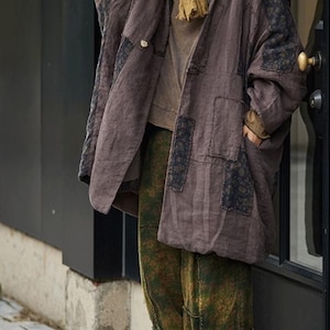 May include: A brown, oversized coat with black patches and a button closure. The coat is paired with olive green patterned pants and a mustard-colored scarf. The outfit is completed with beige shoes, creating a layered, casual look.