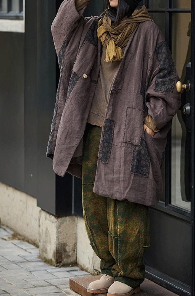 May include: A brown, oversized coat with black patches and a button closure. The coat is paired with olive green patterned pants and a mustard-colored scarf. The outfit is completed with beige shoes, creating a layered, casual look.