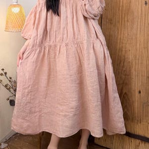 May include: A light pink linen dress with a V-neck and long, puffy sleeves. The dress has a tiered skirt and is knee-length. The person is wearing brown slip-on shoes. A woven lamp and wooden door are in the background.