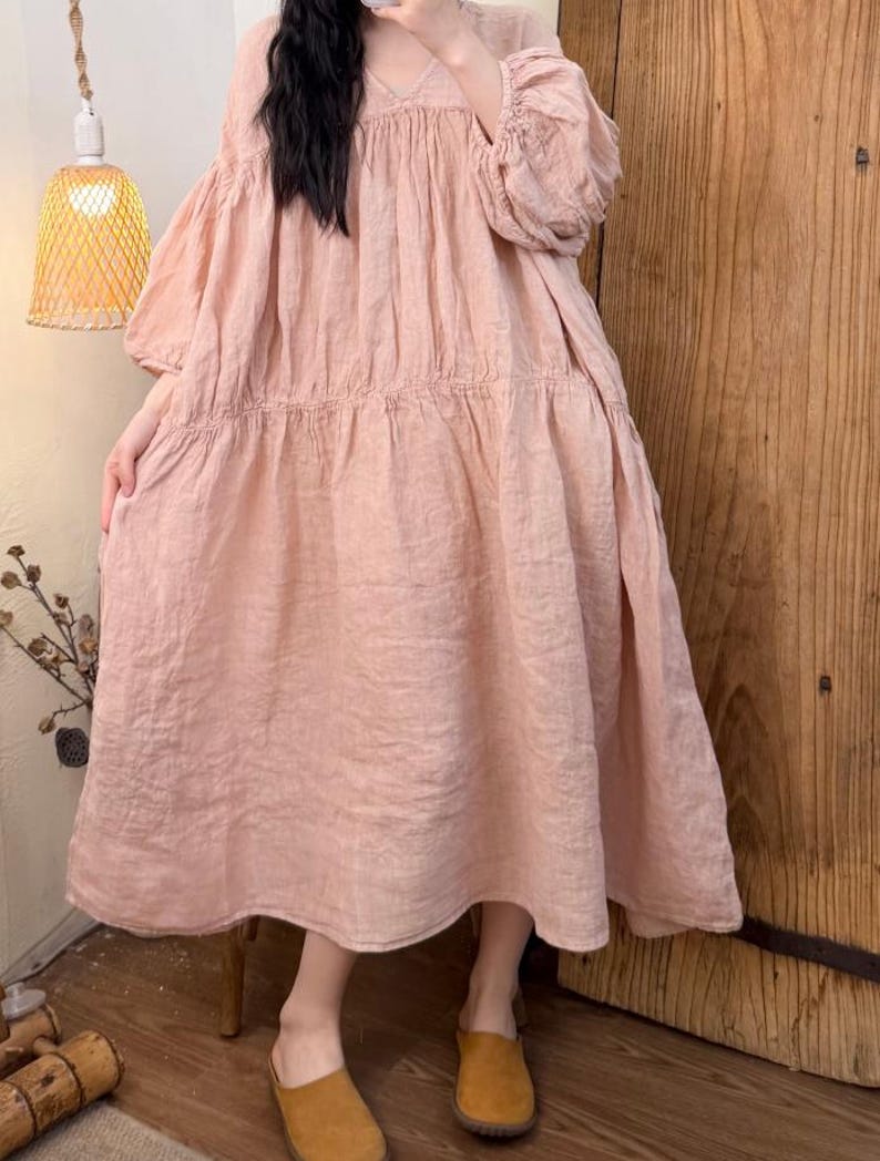 May include: A light pink linen dress with a V-neck and long, puffy sleeves. The dress has a tiered skirt and is knee-length. The person is wearing brown slip-on shoes. A woven lamp and wooden door are in the background.