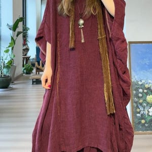 May include: A burgundy-colored dress with a flowing, layered design. The dress features wide sleeves and a draped neckline. A long, brown scarf and a silver pendant necklace add detail. The dress appears to be made of a soft, textured fabric.