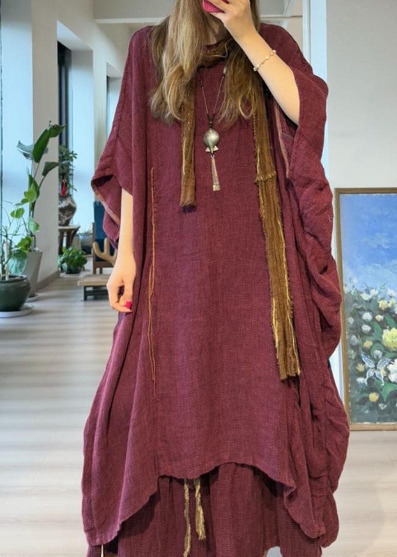 May include: A burgundy-colored dress with a flowing, layered design. The dress features wide sleeves and a draped neckline. A long, brown scarf and a silver pendant necklace add detail. The dress appears to be made of a soft, textured fabric.