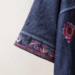 May include: Close-up of a denim garment with embroidered floral and dragon designs. The sleeve features a detailed border with colorful embroidery. The garment is a deep blue color.