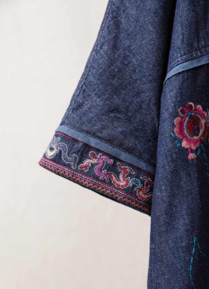 May include: Close-up of a denim garment with embroidered floral and dragon designs. The sleeve features a detailed border with colorful embroidery. The garment is a deep blue color.