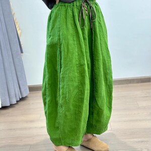 May include: Bright green linen pants with a drawstring waist. The pants have a loose, flowing design and a slightly gathered hem. The fabric appears to be textured. The pants are paired with tan colored slippers.