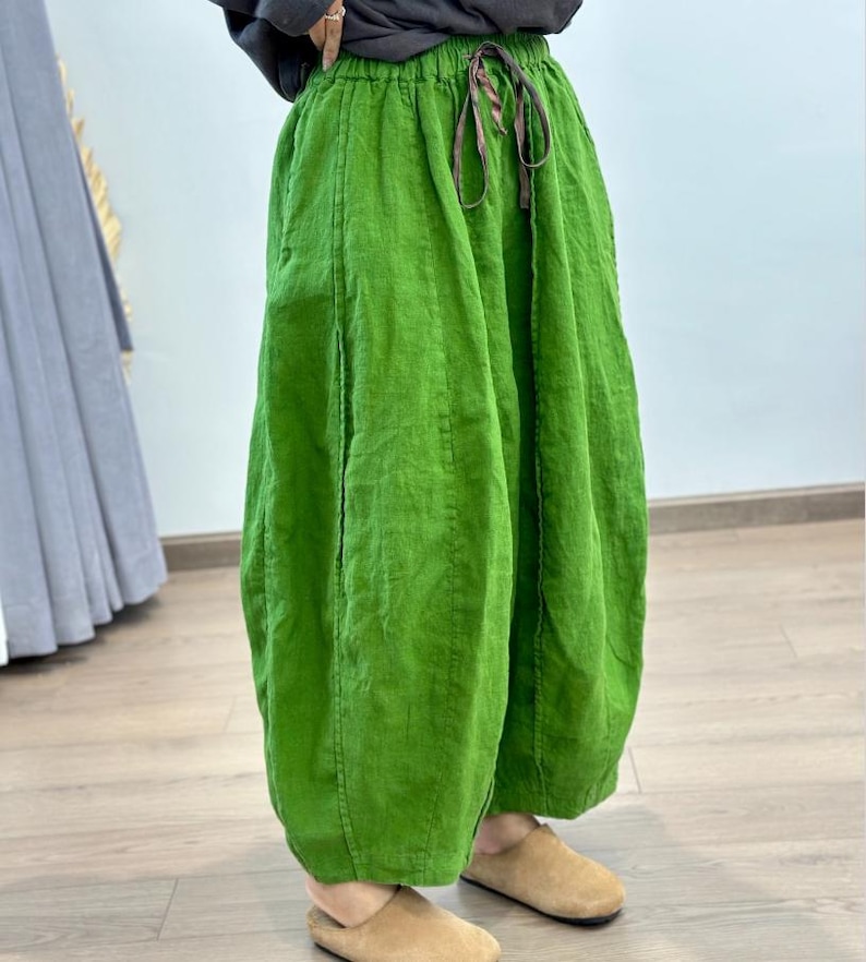 May include: Bright green linen pants with a drawstring waist. The pants have a loose, flowing design and a slightly gathered hem. The fabric appears to be textured. The pants are paired with tan colored slippers.