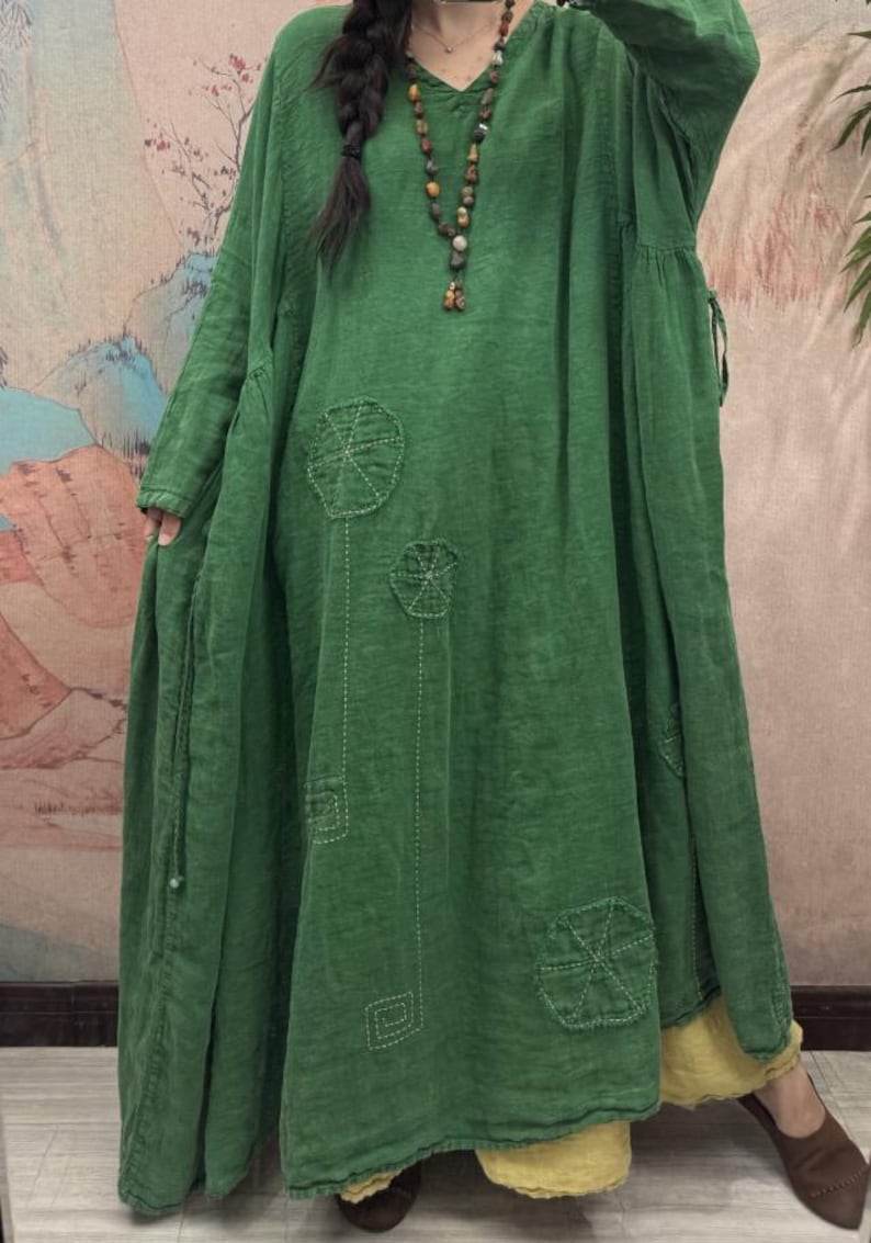 May include: A long, flowing green dress with a V-neck and long sleeves. The dress features embroidered circular and square designs. The model is wearing a beaded necklace. The dress is layered over a yellow garment.