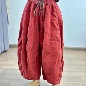 May include: Red linen pants with a drawstring waist and wide, voluminous legs. The pants have a unique, gathered design and are made of a textured fabric. The pants are paired with tan slip-on shoes.