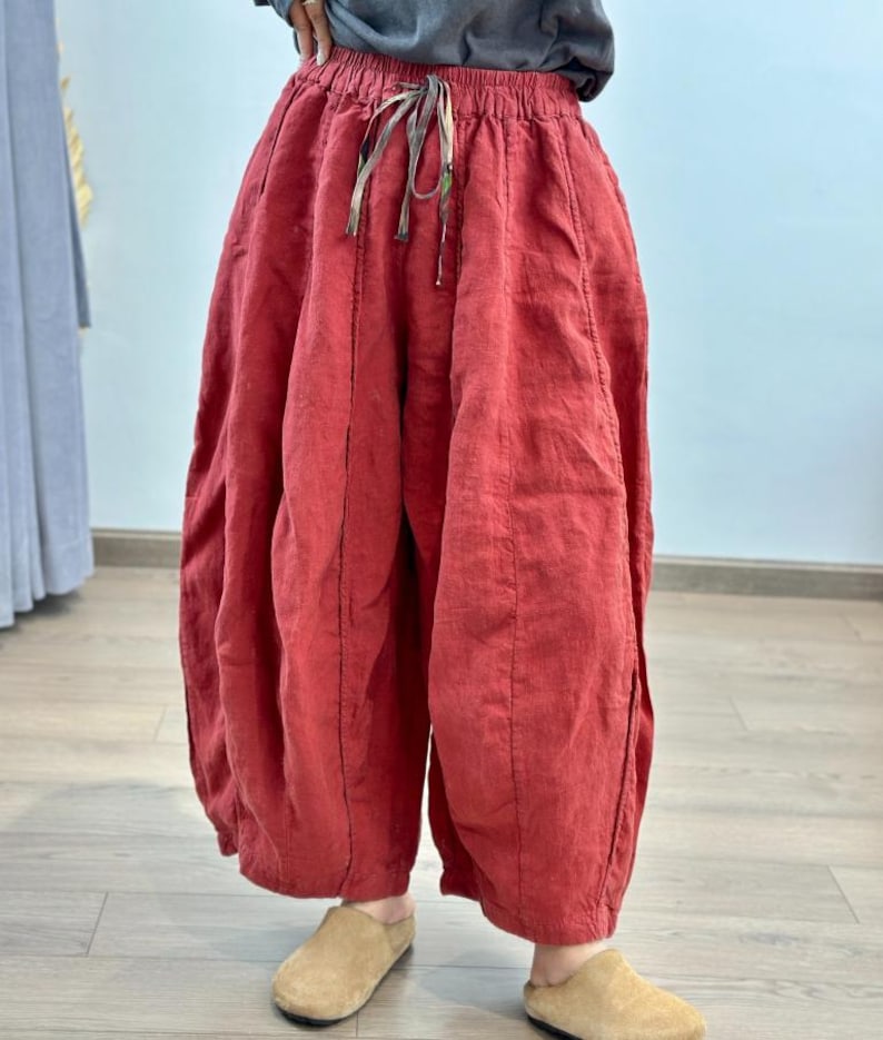 May include: Red linen pants with a drawstring waist and wide, voluminous legs. The pants have a unique, gathered design and are made of a textured fabric. The pants are paired with tan slip-on shoes.