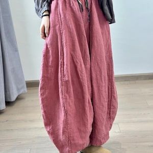 May include: Loose-fitting, dusty rose-colored linen pants with a drawstring waist. The pants have a unique, voluminous shape, gathered at the ankles. The model is wearing tan-colored slip-on shoes. The pants are made of a lightweight fabric.