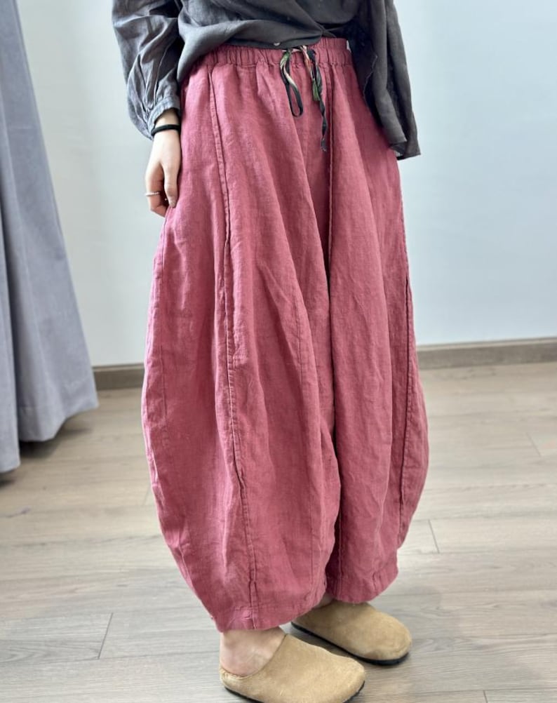 May include: Loose-fitting, dusty rose-colored linen pants with a drawstring waist. The pants have a unique, voluminous shape, gathered at the ankles. The model is wearing tan-colored slip-on shoes. The pants are made of a lightweight fabric.
