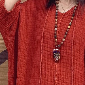 May include: A red linen tunic top with a long, beaded necklace featuring a small, colorful pendant. The top has a relaxed fit and a V-neckline.